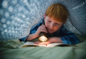 Boy (8-9) reading book under bed covers --- Image by © Tetra Images/Corbis